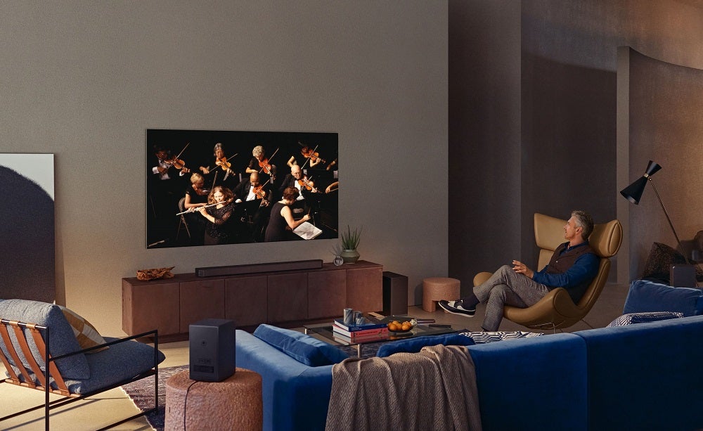 Un téléviseur noir Samsung Neo QLED 4K accroché à un mur avec un vieil homme assis sur une chaise et regardant la télévision