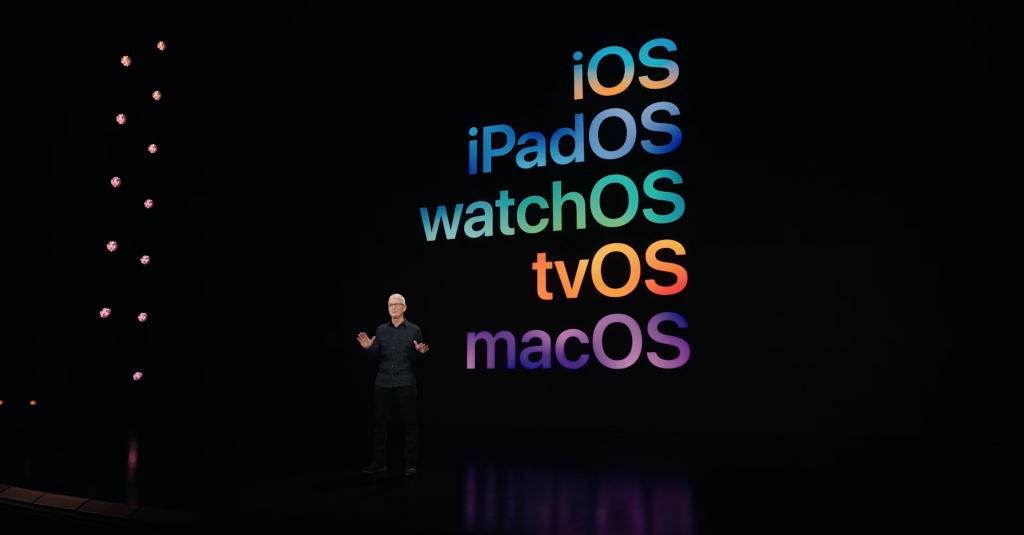 Tim Cook debout sur scène dans une tenue noire avec iOS, iPadOS, watchOS, tvOS et macOS écrits sur l'écran derrière lui