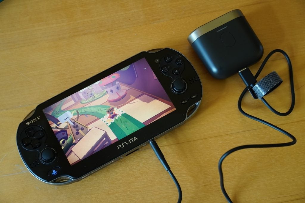 Une Playstation PS Vita noire de Sony affichant un jeu avec des commandes de jeu de chaque côté, une prise jack connectée et l'autre extrémité connectée à un haut-parleur, présentant la transmission audio Bowers et Wilkins P17